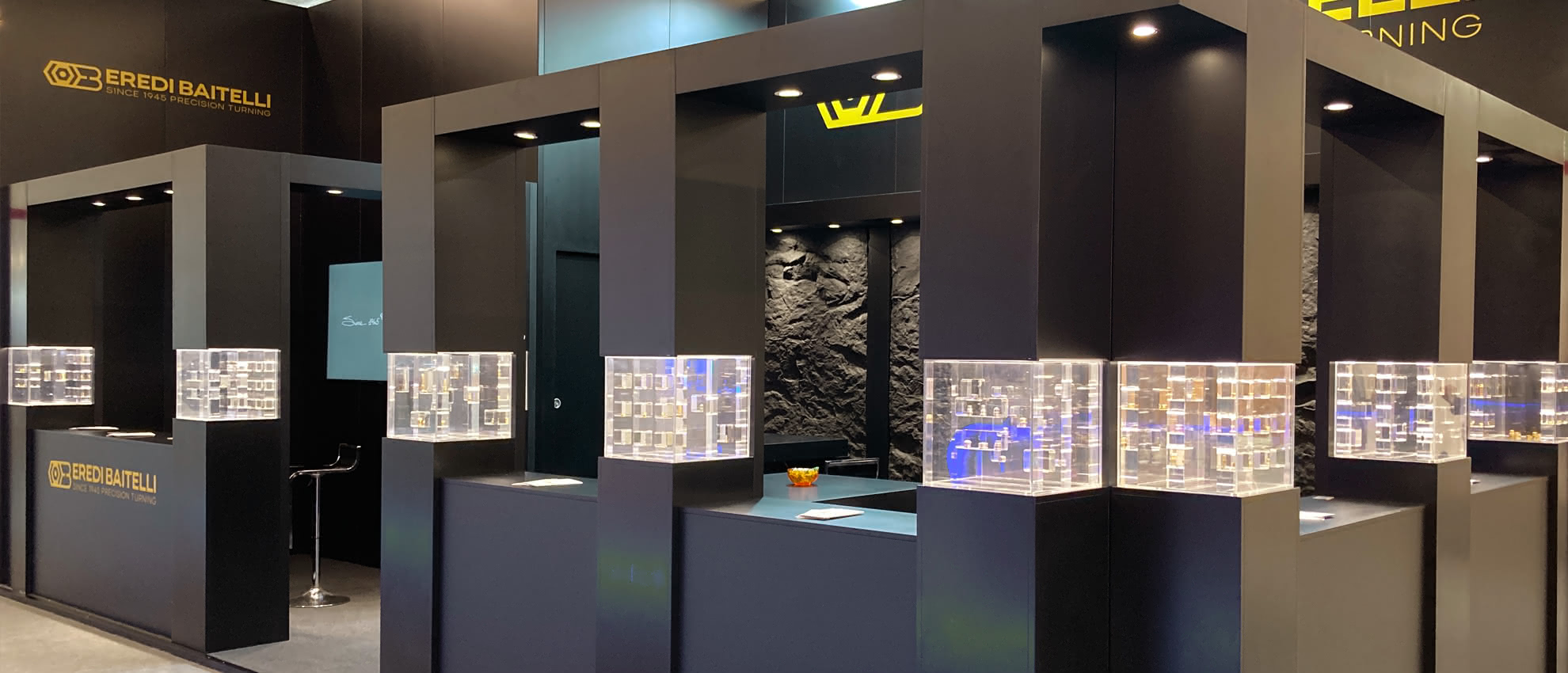 Seitenansicht des Eredi Baitelli Messestandes mit modularen schwarzen Elementen und transparenten Vitrinen in Inselanordnung.